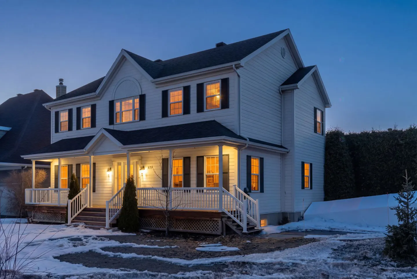 Two-story white house with black shutters and a porch, lit by warm lights at dusk. Snow is on the ground.