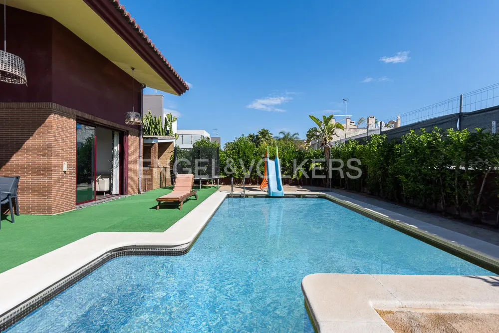 A backyard with a pool, slide, and lounge chair on artificial grass. The house is brick with a brown roof. The sky is blue.