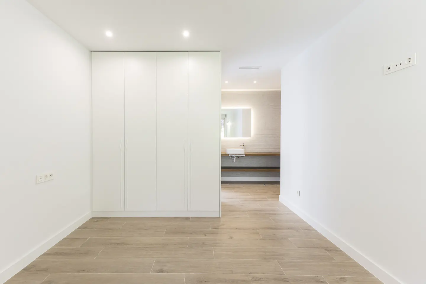 Minimalist room with white walls, light wood floors, a large white wardrobe, and a view into a modern bathroom.