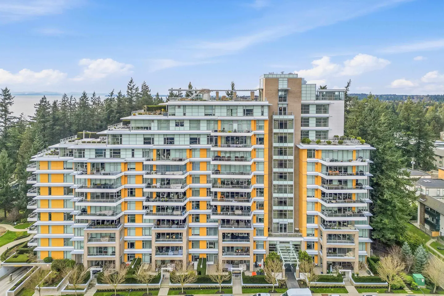 Modern condo building with yellow accents and balconies, surrounded by trees under a blue sky.