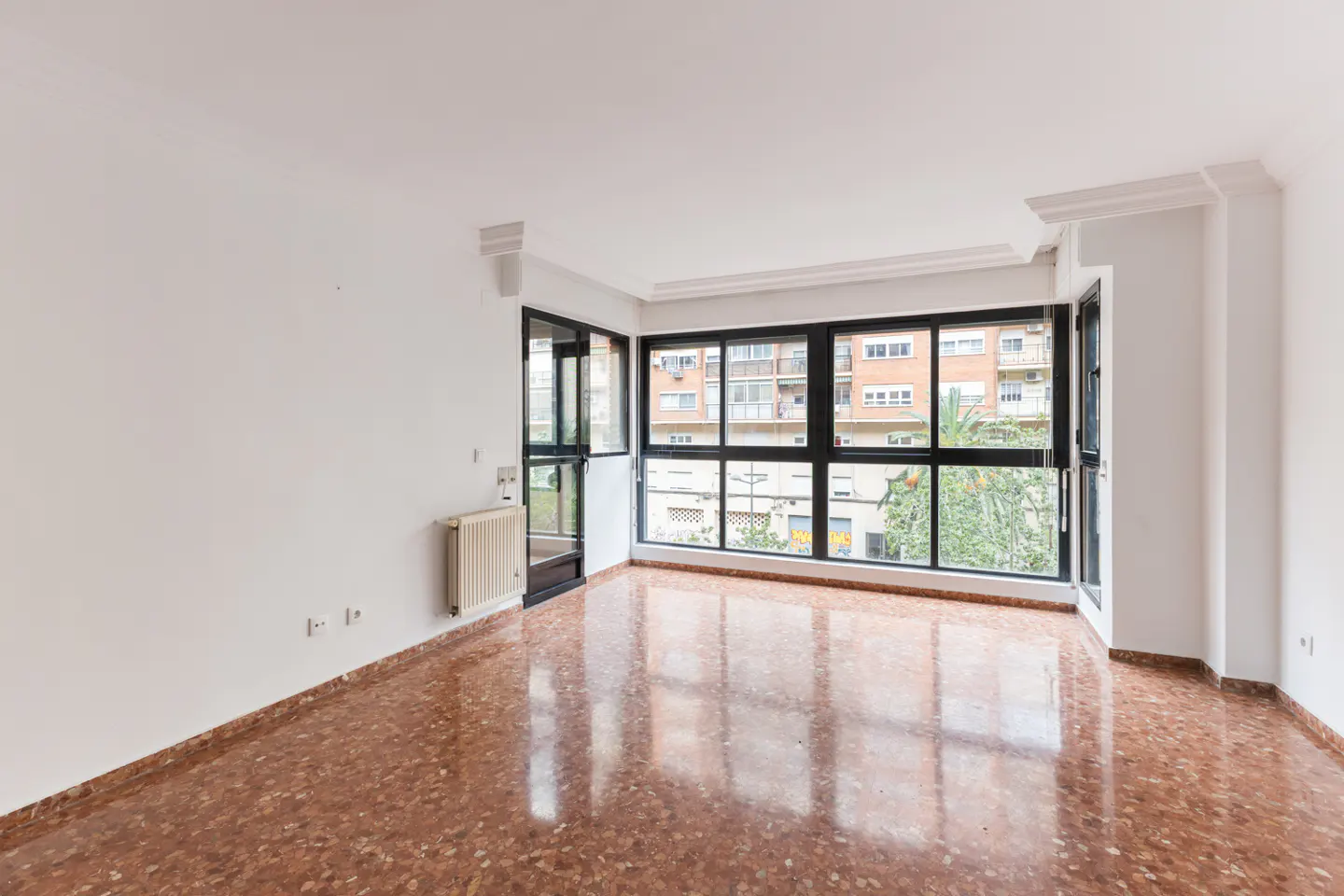 Bright, empty room with white walls, red marble floor, and large black-framed windows overlooking a city street.