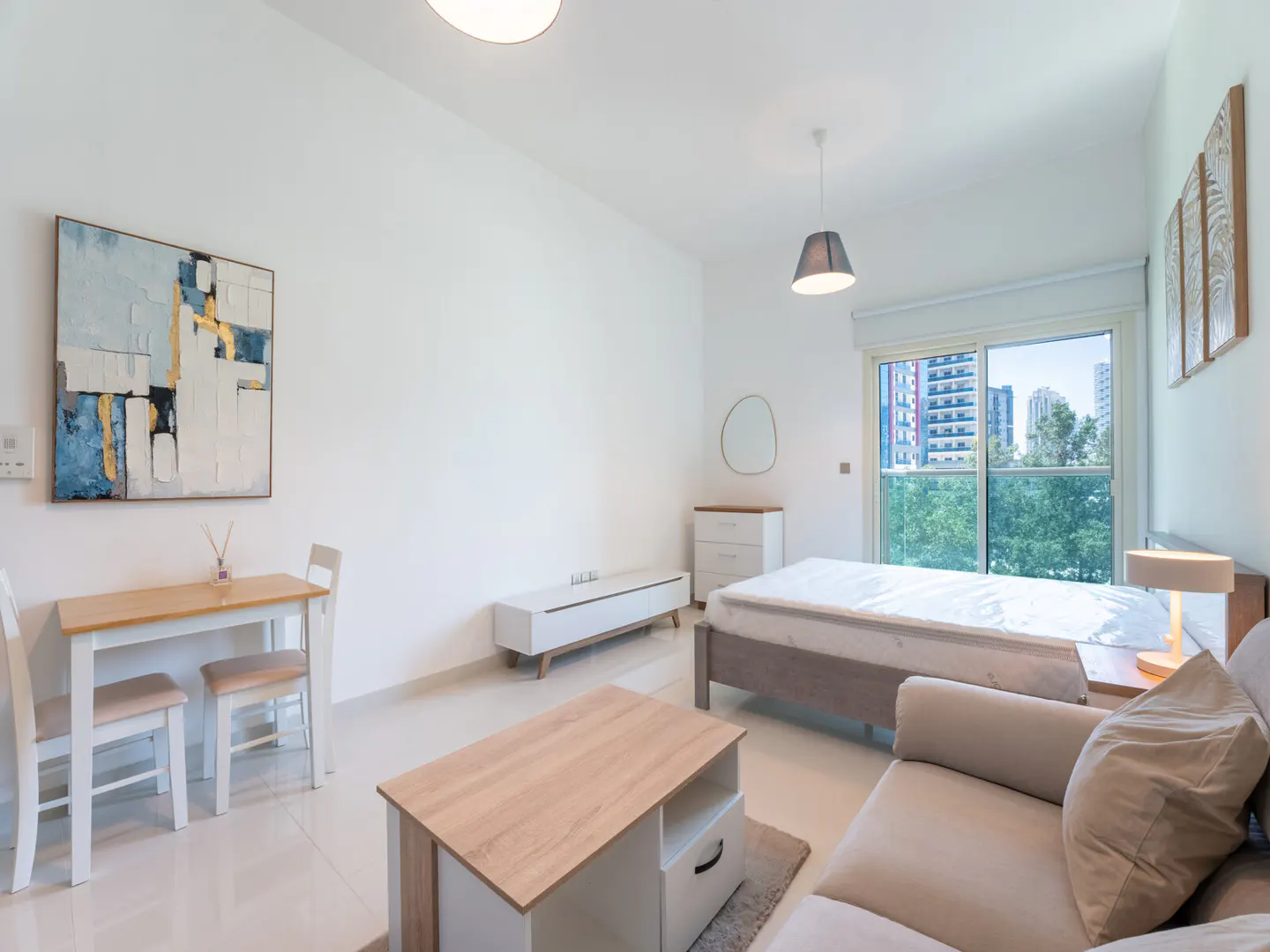 Bright studio apartment with white walls, a bed, sofa, table, and abstract art. A window shows a city view.