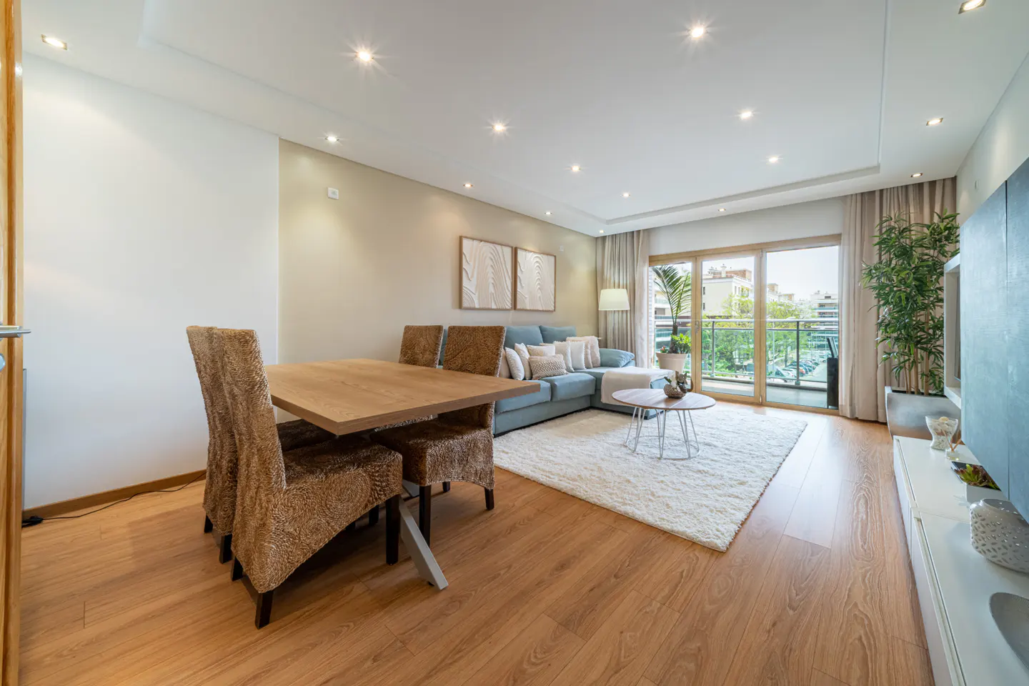 Bright living room with wood floors, a dining table with four chairs, a blue sofa, and a white rug. A sliding glass door leads to a balcony.