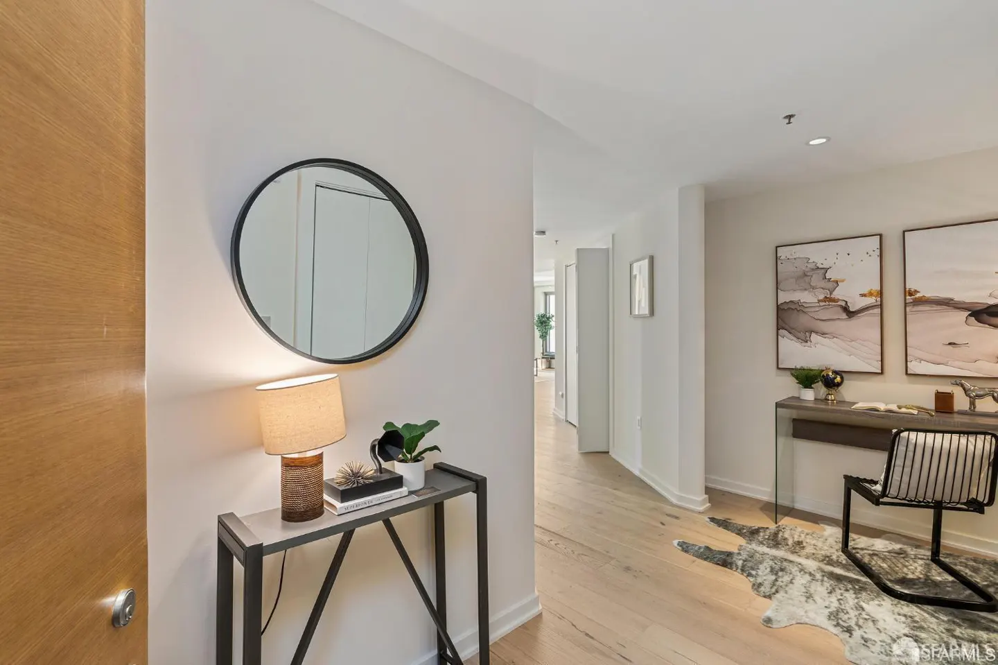 Entryway with light wood floors, white walls, and a black-framed round mirror above a console table with a lamp. Hallway leads to other rooms.