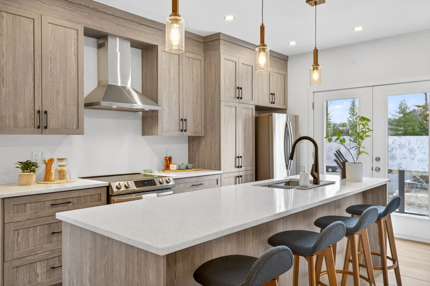 Bright kitchen with wood cabinets, stainless steel appliances, and a white countertop island with four gray stools. Three pendant lights hang above.
