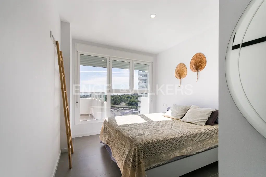 A bright bedroom with a bed, balcony, and decorative ladder. The walls are white, and the bed has a beige cover. Outside, there is a view of trees and water.