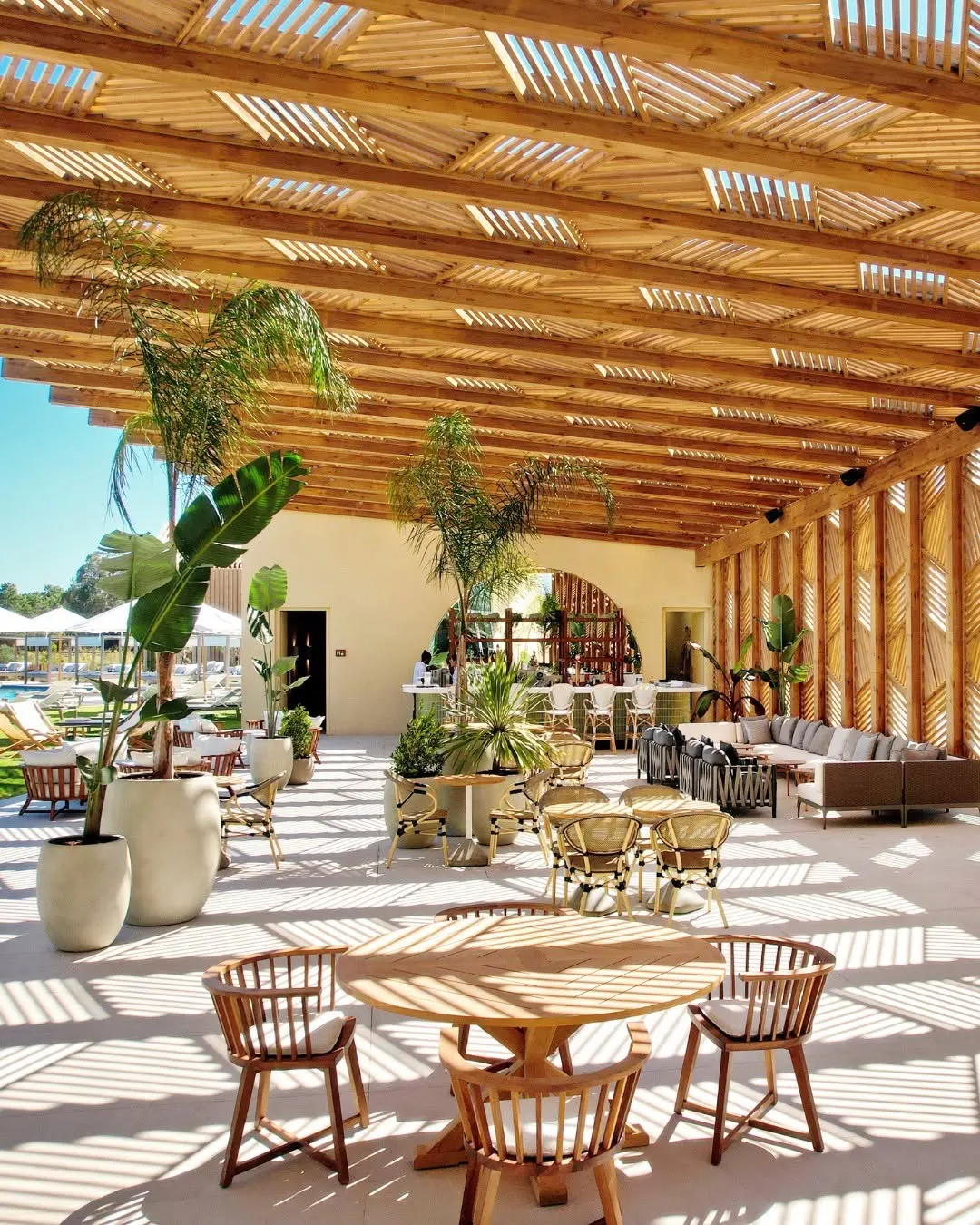 Outdoor patio with wooden tables and chairs under a slatted wooden roof, casting shadows. Plants in pots, a bar, and lounge seating are visible.