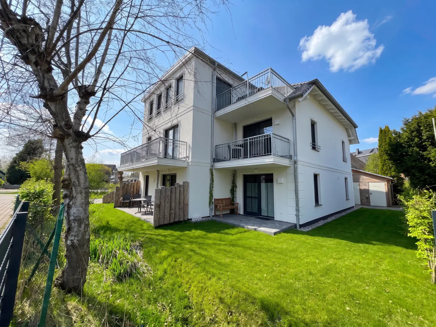 A three-story white building with balconies, set on a green lawn under a blue sky with a few clouds.