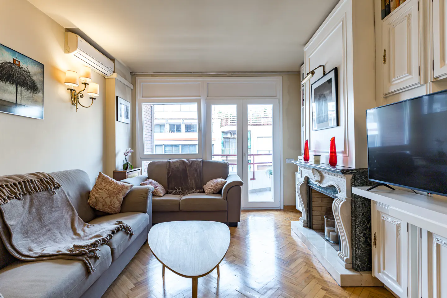 Living room with two sofas, a triangular table, a fireplace, and a TV on a white cabinet. The floor is wooden parquet.