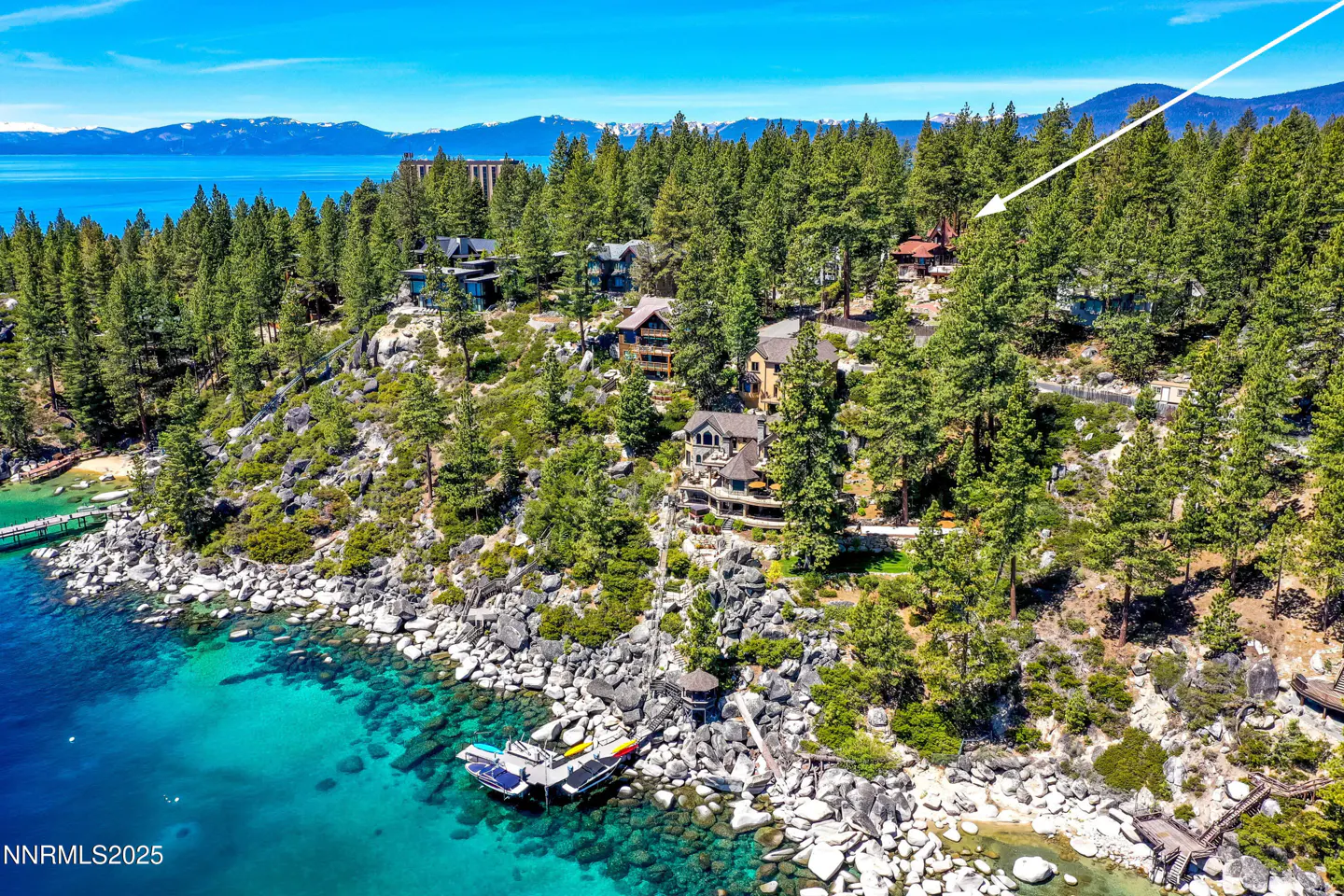 Aerial view of Lake Tahoe shoreline with turquoise water, rocky beach, pier, boats, and homes nestled among green trees. Mountains in the distance.