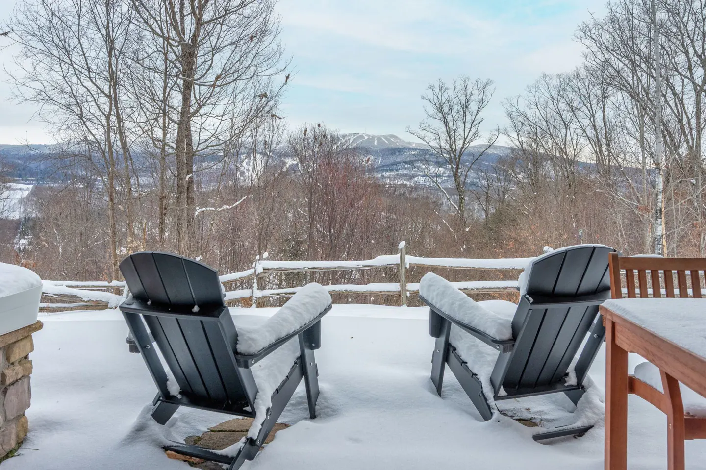 Snowy patio with two black Adirondack chairs facing a mountain view. A wooden fence and table are partially covered in snow.
