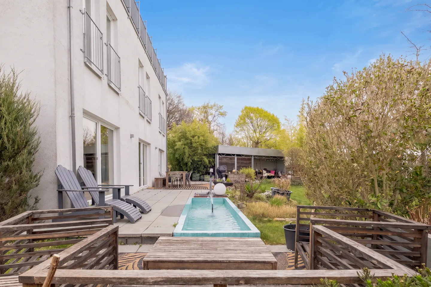 A backyard with a rectangular fountain, chairs, and a white building under a blue sky.