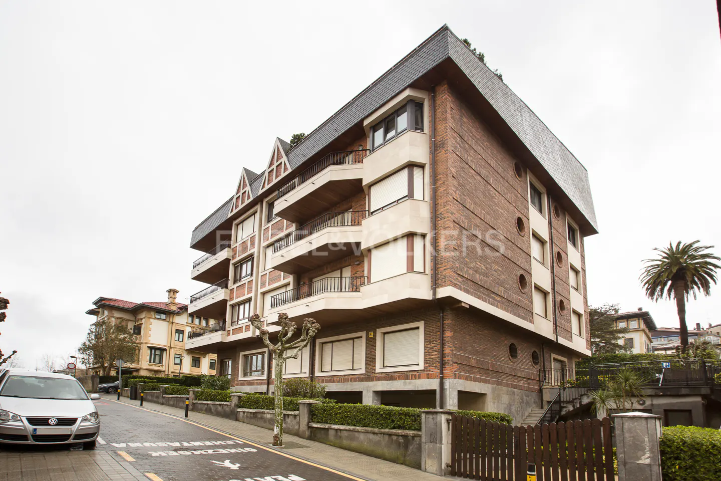Exterior view of a brick apartment building with balconies, a gray roof, and a silver car parked on the street.