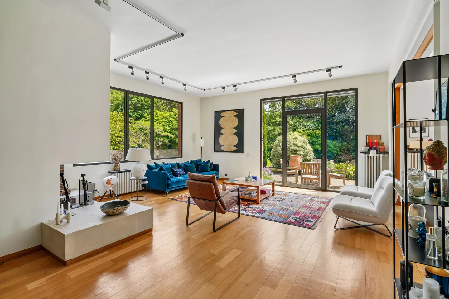 Modern living room with hardwood floors, blue sofa, leather chair, white armchairs, and large windows overlooking a garden.