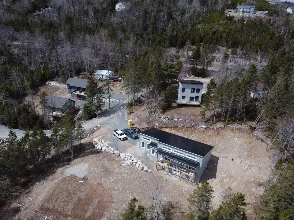 Aerial view of a modern gray house with a black roof, surrounded by trees and other houses. Two cars are parked in front.