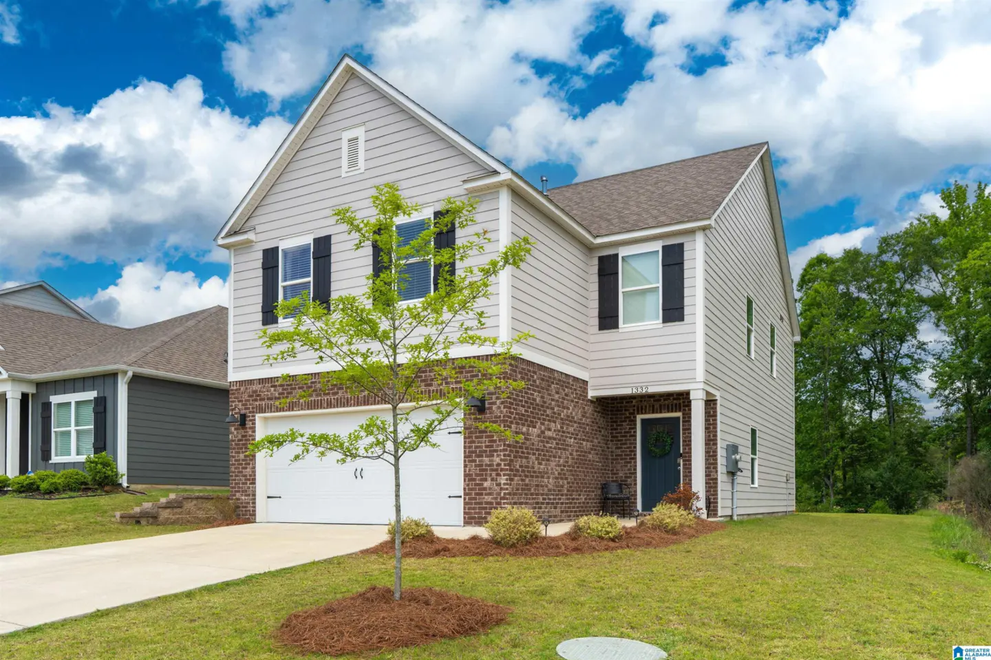 Two-story suburban home with light gray siding, brick accents, white garage, and black shutters under a blue cloudy sky.