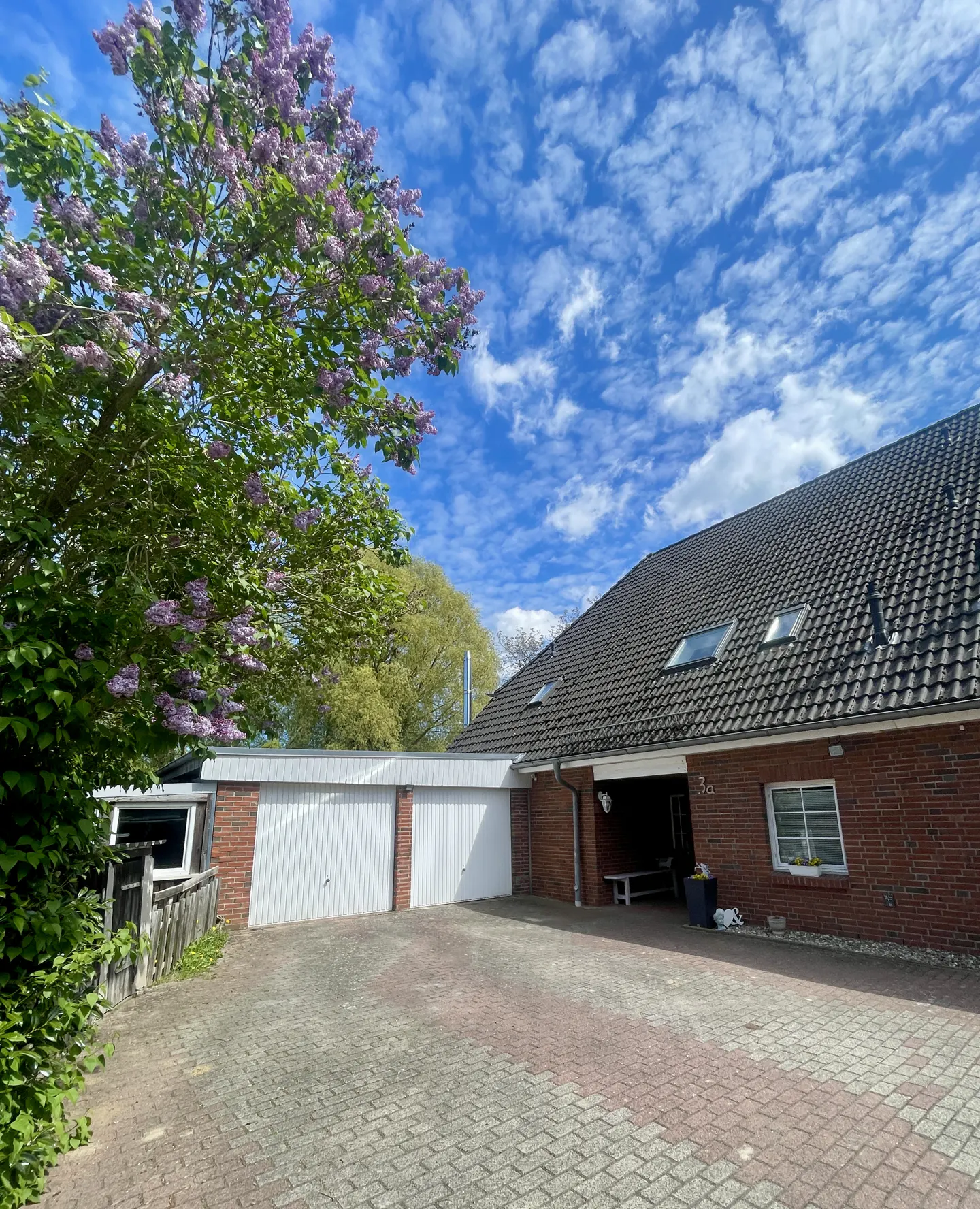 Brick house with a gray roof, white garage doors, and a lilac tree against a blue sky.