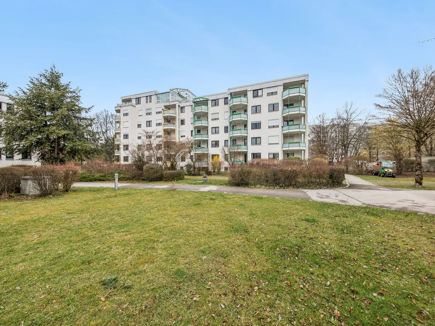 Exterior view of a white apartment building with green balconies, surrounded by a green lawn and trees under a blue sky.