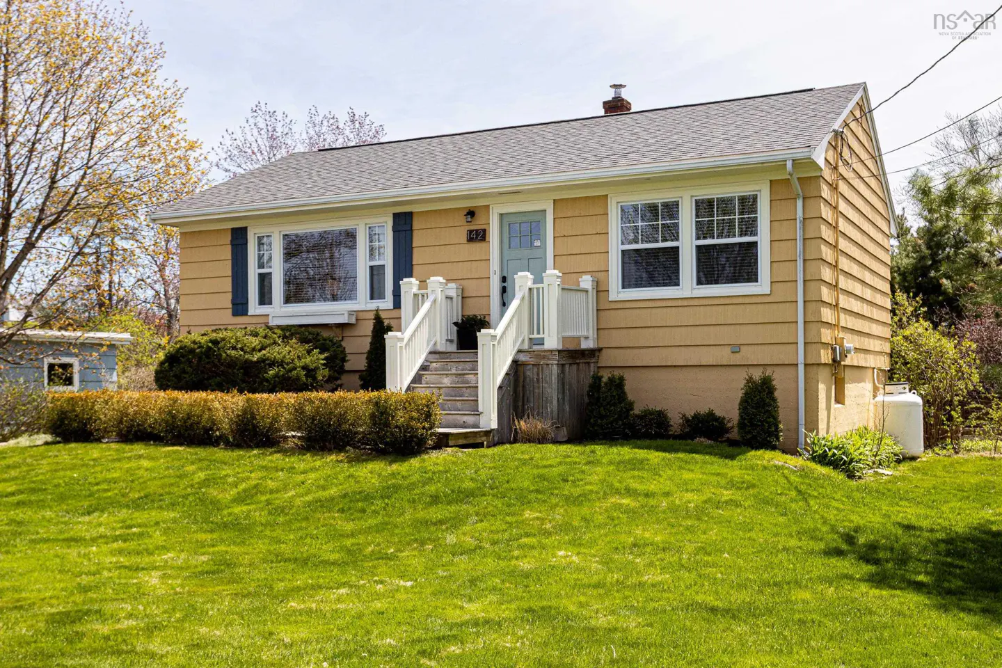 Tan house with a light blue door and white trim, with a green lawn in front. Bushes line the front of the house.