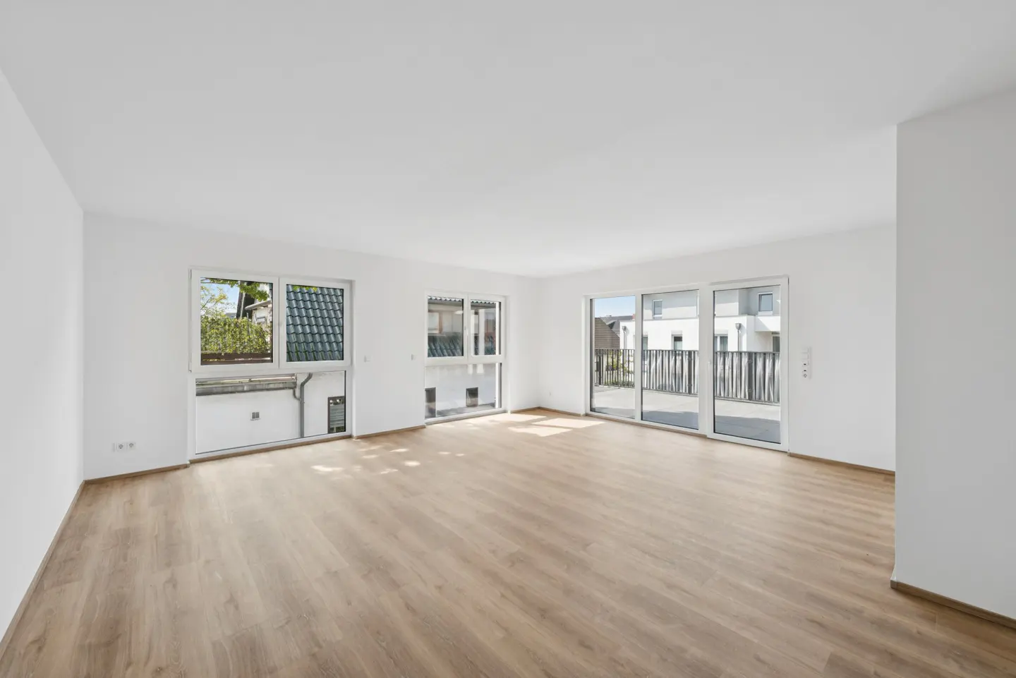 Bright, empty room with light wood floors, white walls, and large windows. Outside, green trees and buildings are visible.