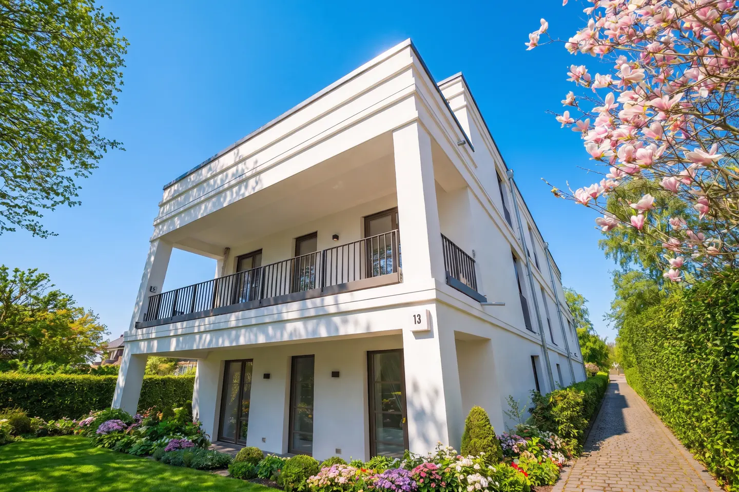 A low-angle view of a modern, white, two-story house with a balcony, green lawn, and colorful flowers.