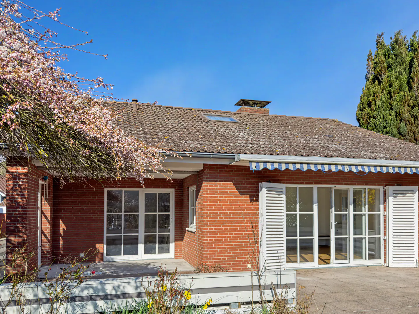 Red brick house with white trim, a brown tile roof, and a blue and white striped awning on a sunny day.