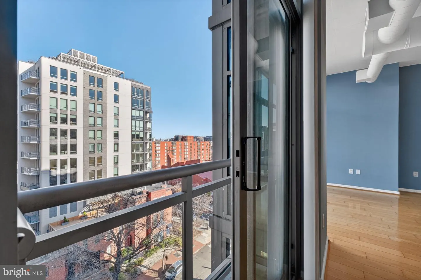 View from a condo balcony with a metal railing, overlooking city buildings and a street. Inside, a blue wall and wood floors are visible.