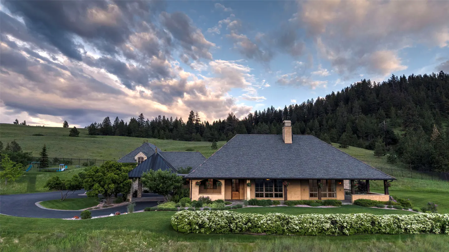 A tan house with a dark roof sits on a green lawn before a forested hillside under a dramatic, cloudy sunset sky.