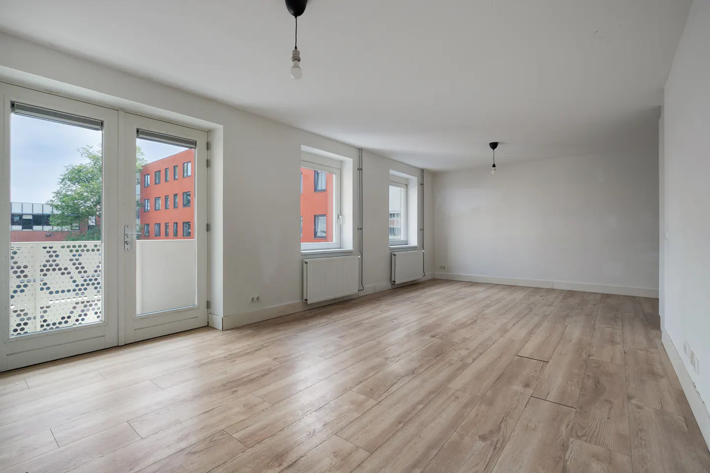 Bright, empty room with light wood floors, white walls, and two windows. A glass door leads to a balcony.