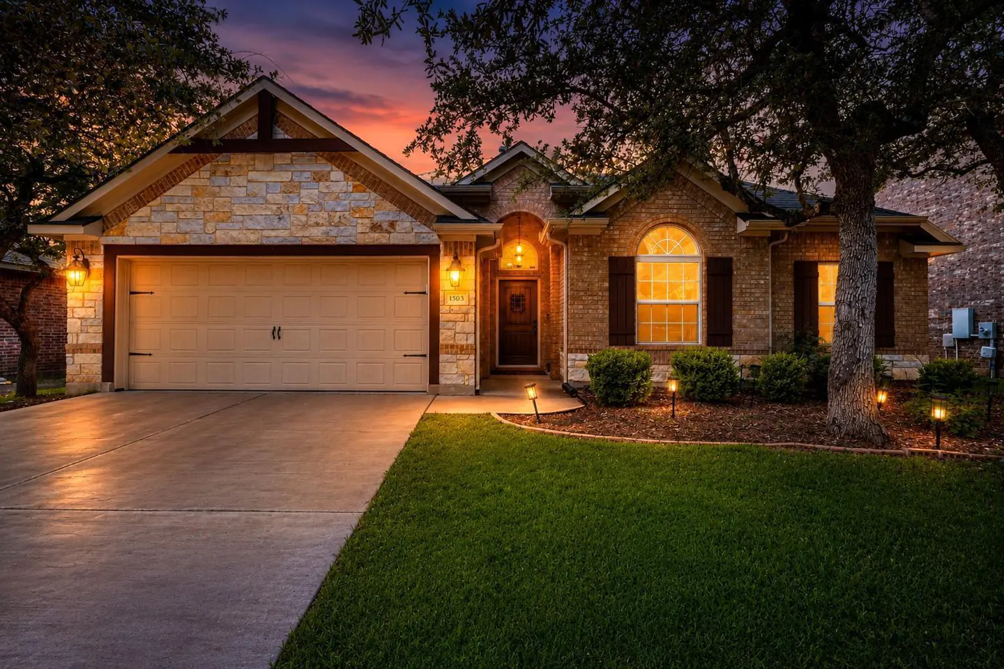 Exterior shot of a single-story home with a stone and brick facade, a beige garage door, and a green lawn at dusk.
