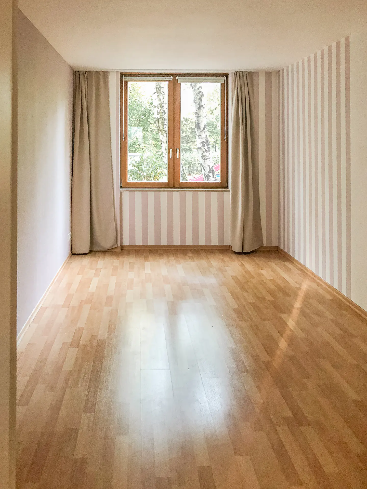 Empty room with wood floor, striped wallpaper, and a window with beige curtains.