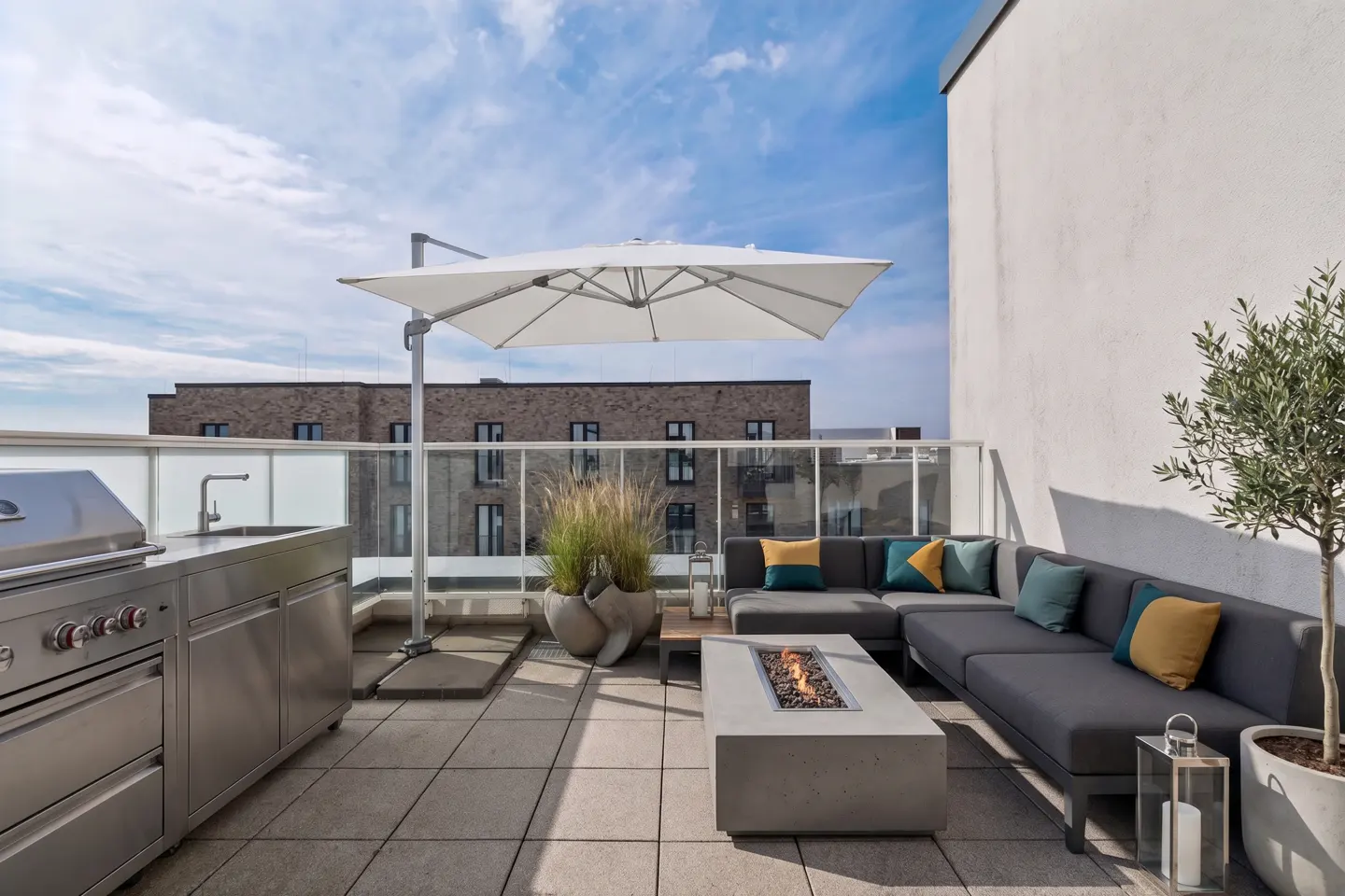 A modern rooftop terrace with gray sectional seating, a fire pit, and an outdoor kitchen under a white umbrella.