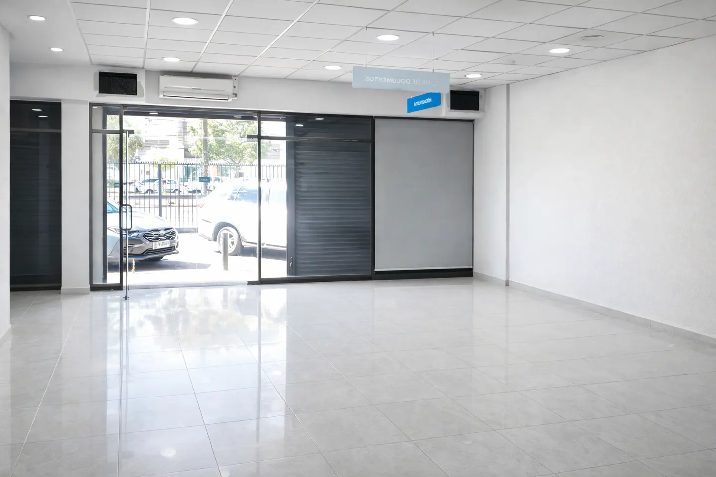 Bright, empty commercial space with white walls, gray tile floor, and glass doors to the street. A gray roller blind is partially lowered.