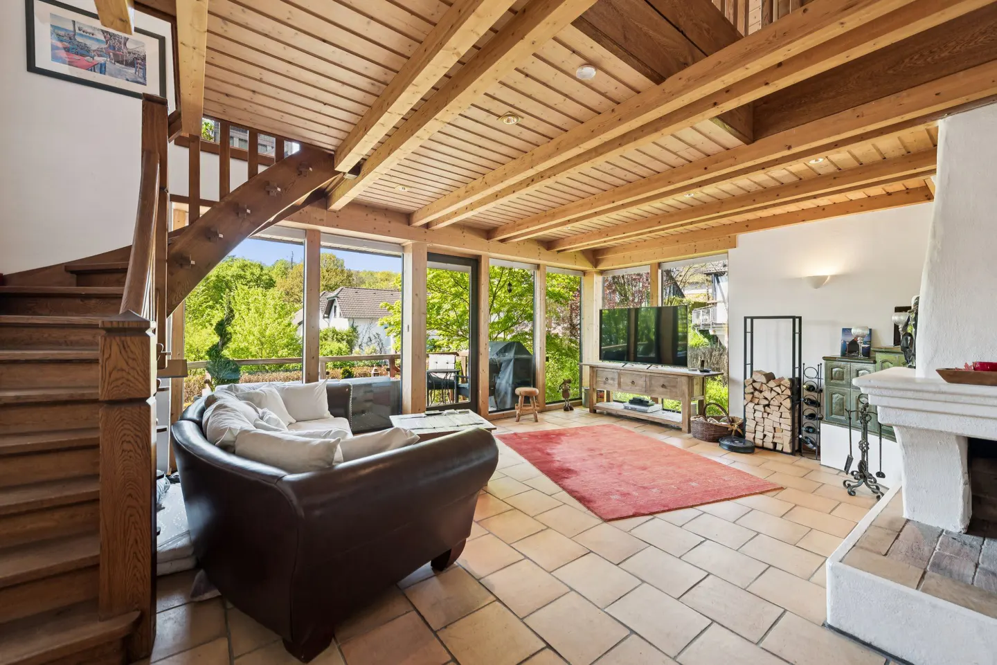 Living room with wood-beamed ceiling, stone floors, and large windows. Brown leather sofa, red rug, and white fireplace. Stairs to the left.