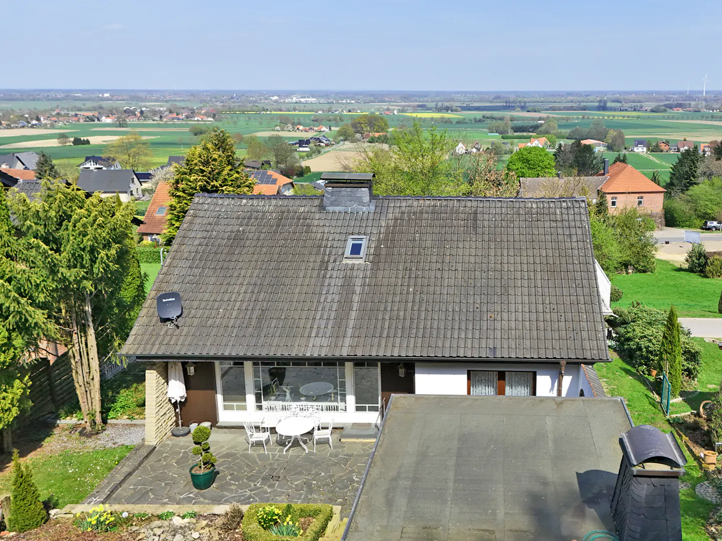 Aerial view of a one-story house with a gray tile roof, stone patio, and white outdoor furniture, overlooking a green landscape.