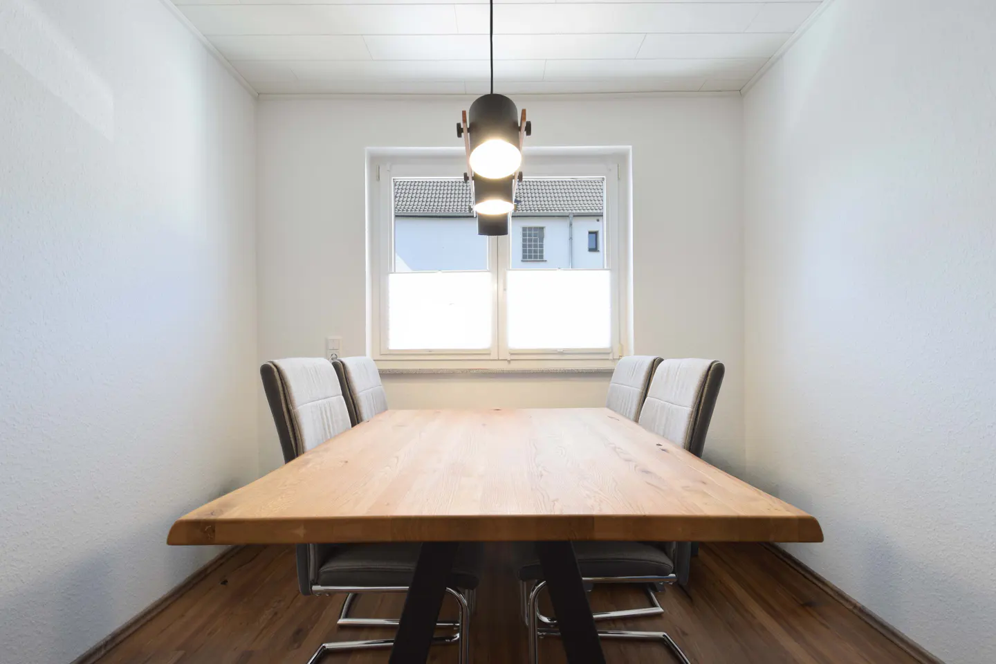 Dining room with a light wood table, four gray chairs, and a modern black pendant light. White walls and wood floors.