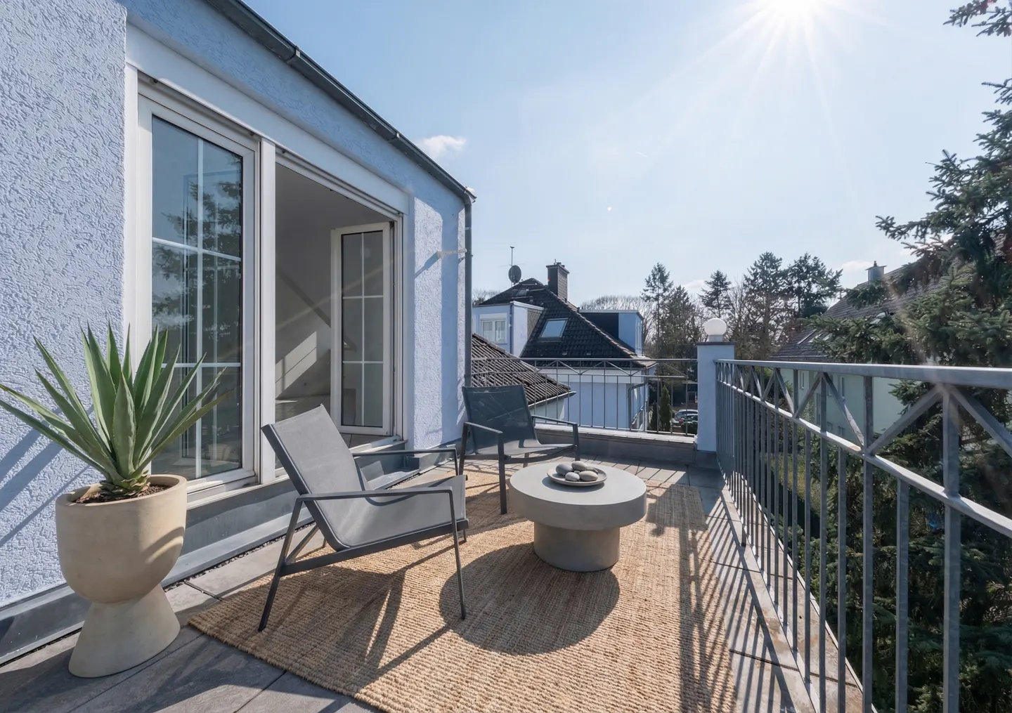 A sunlit balcony with two gray chairs, a round table, and a potted plant on a woven rug. Railing overlooks houses and trees.