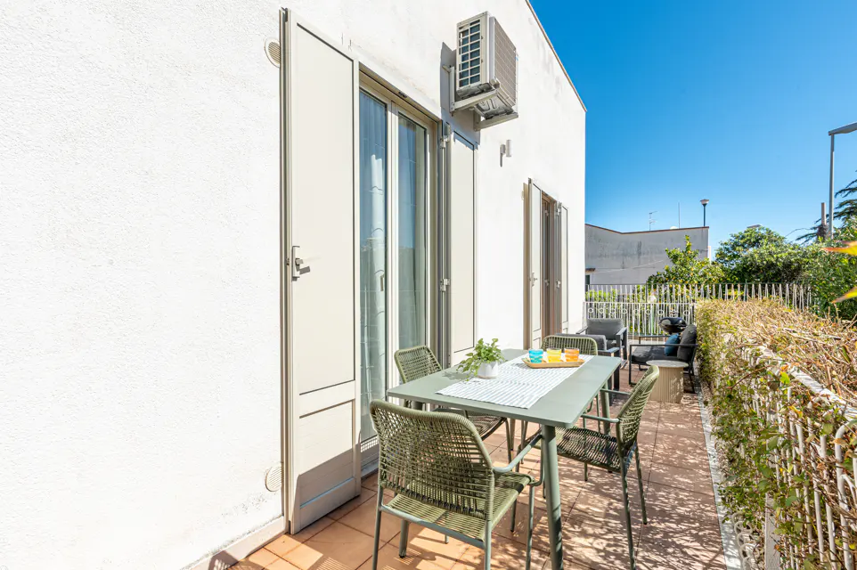 Outdoor patio with a green table and four chairs. A white building with a sliding glass door and an air conditioner is visible. The patio has a hedge and a view of the neighborhood.