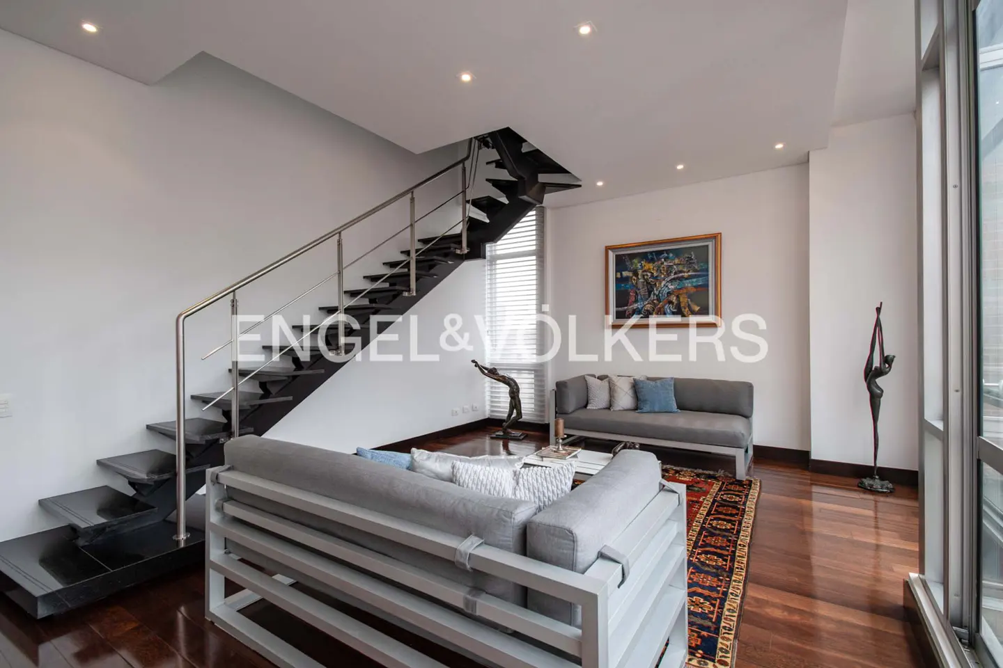 Living room with gray sofas, wood floors, and black stairs with stainless steel railing. Artwork and sculptures decorate the space.