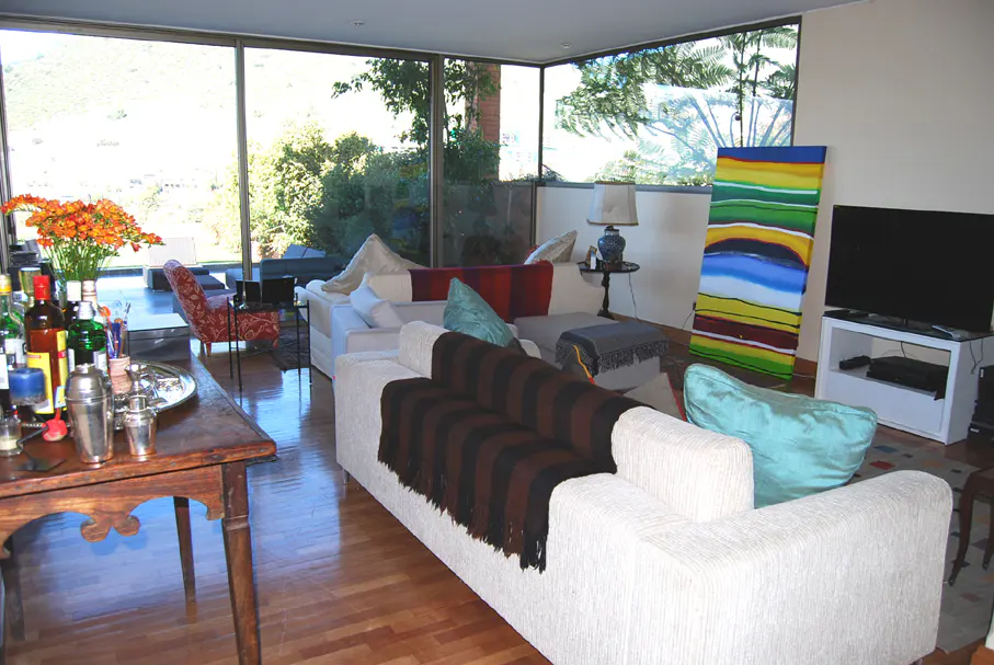Living room with white sofas, wood floors, and floor-to-ceiling windows overlooking a green landscape. A bar cart with drinks sits on a wooden table.