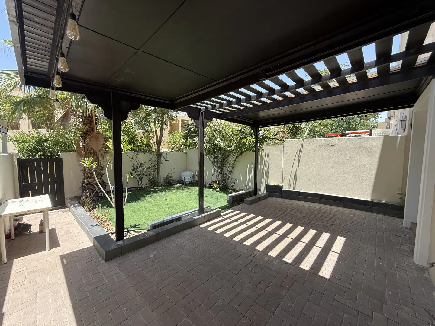 Outdoor patio with a dark pergola casting shadows on the brick floor. Green lawn and trees are visible in the background.
