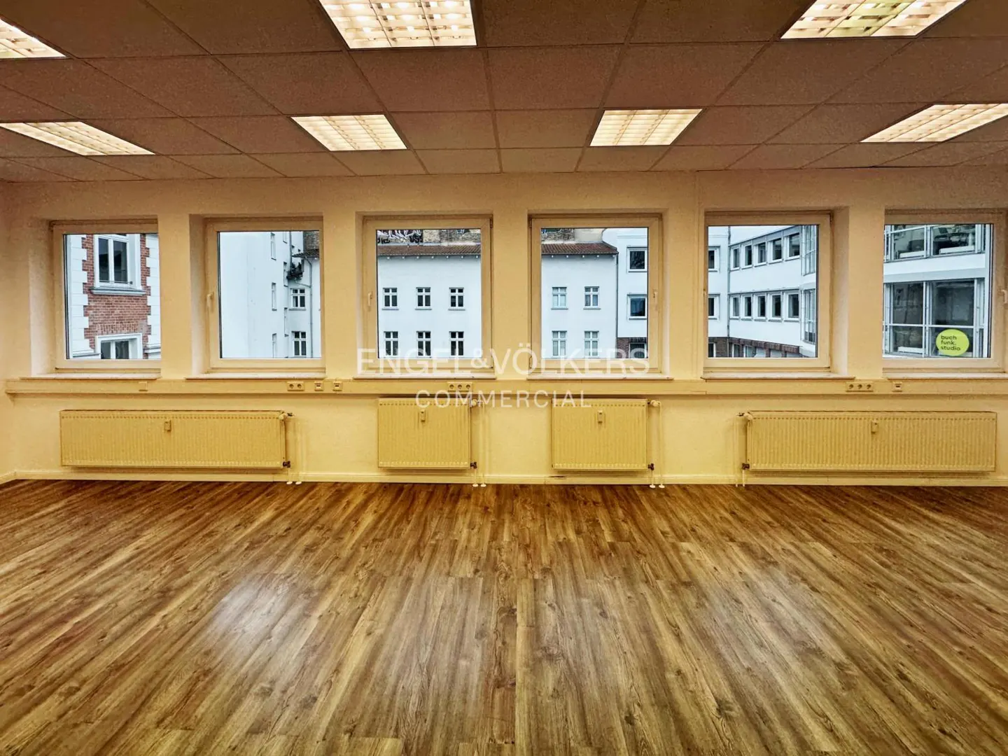 Empty office space with wood floors, white walls, and five windows showing city buildings. Radiators are under the windows.