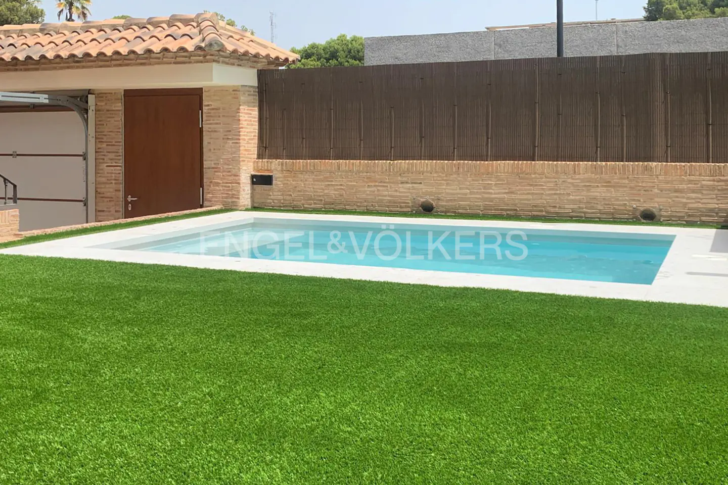 A rectangular pool with blue water is surrounded by green grass and a brick wall with a brown fence.