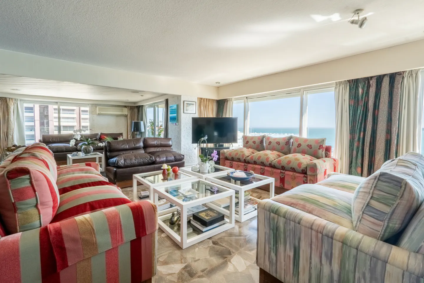 Bright living room with striped sofas, leather couches, and white square coffee tables. Ocean view through large windows.