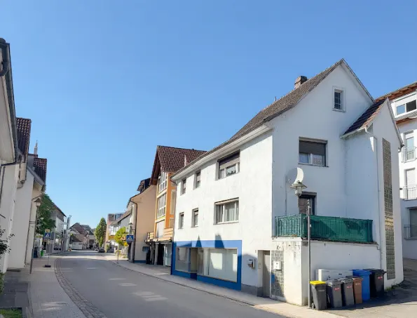 Street view of white buildings with red roofs under a clear blue sky. Trash bins line the sidewalk.