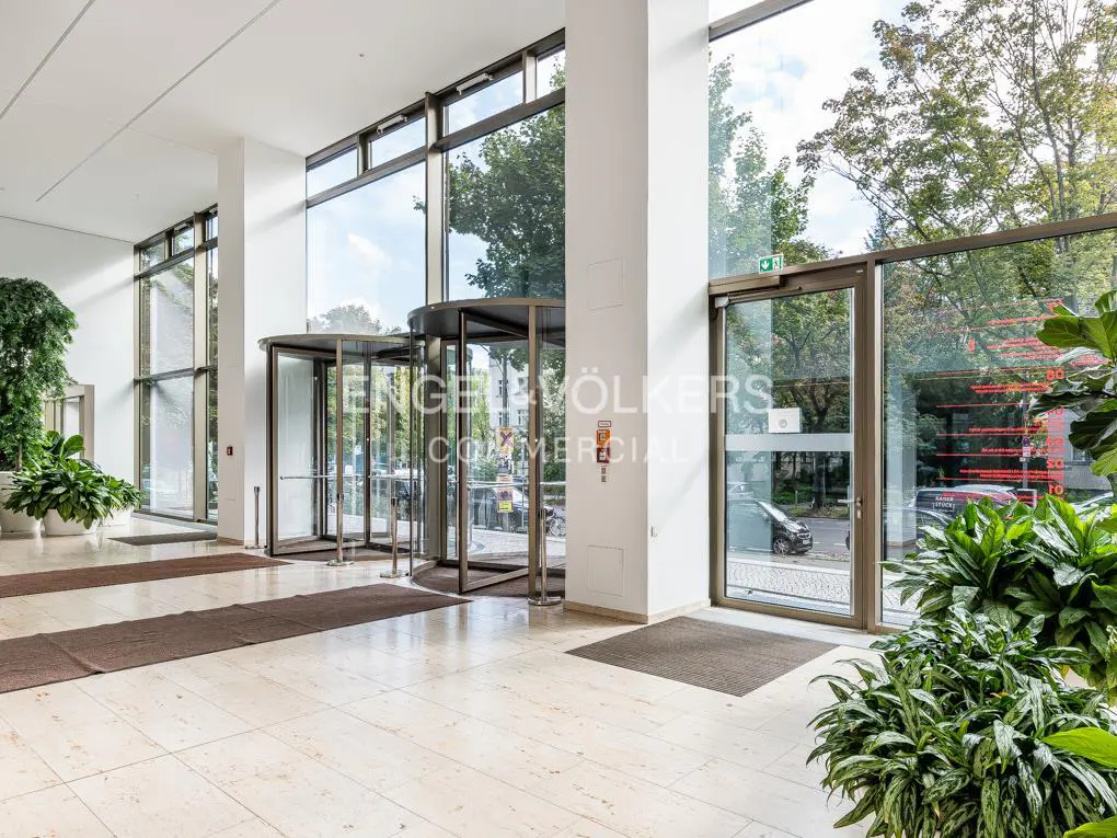Bright lobby with revolving doors, large windows, and potted plants. Beige tile floor and brown mats. Engel & Völkers logo visible.
