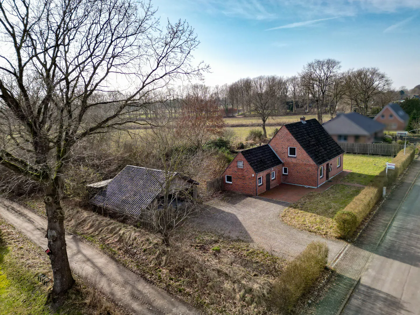 Aerial view of a red brick house with a black roof, a gravel driveway, and a shed with a gray roof. A large tree is on the left.