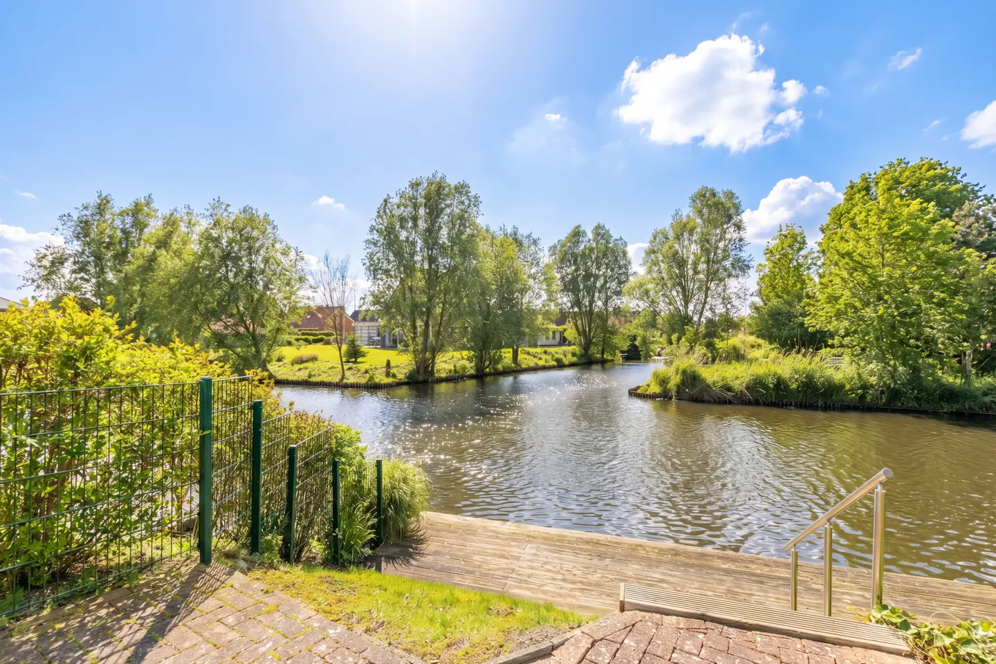 Waterfront property view with a canal, trees, and blue sky. A wooden deck with metal stairs leads to the water. Green fence and brick patio in the foreground.
