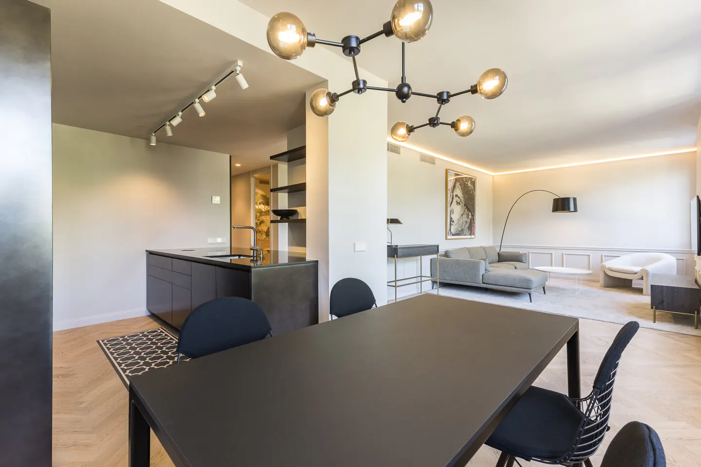 Open-concept living space with a black dining table, chairs, and a modern chandelier. Kitchen and living room visible in the background.