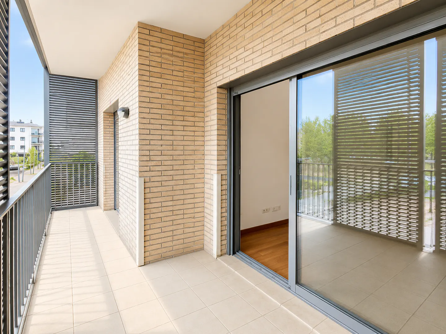 Modern balcony with light brick walls, tiled floor, metal railings, and large sliding glass doors leading to an interior.